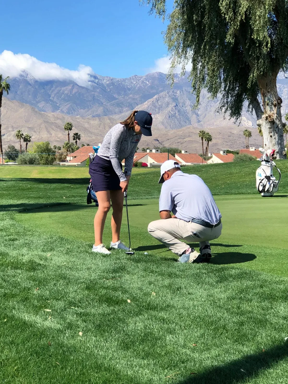 A coach in a light polo crouches to check a putt beside a player, with mountains behind