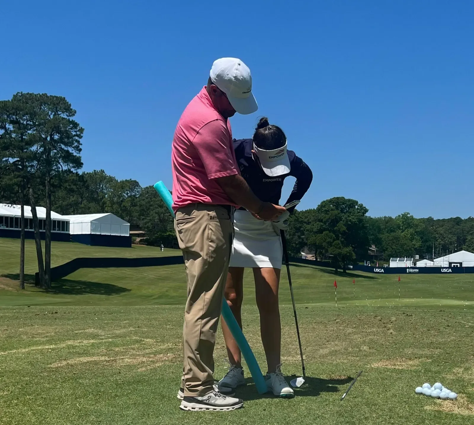 A coach in a pink polo stands behind a player, guiding her hands on the club at a range