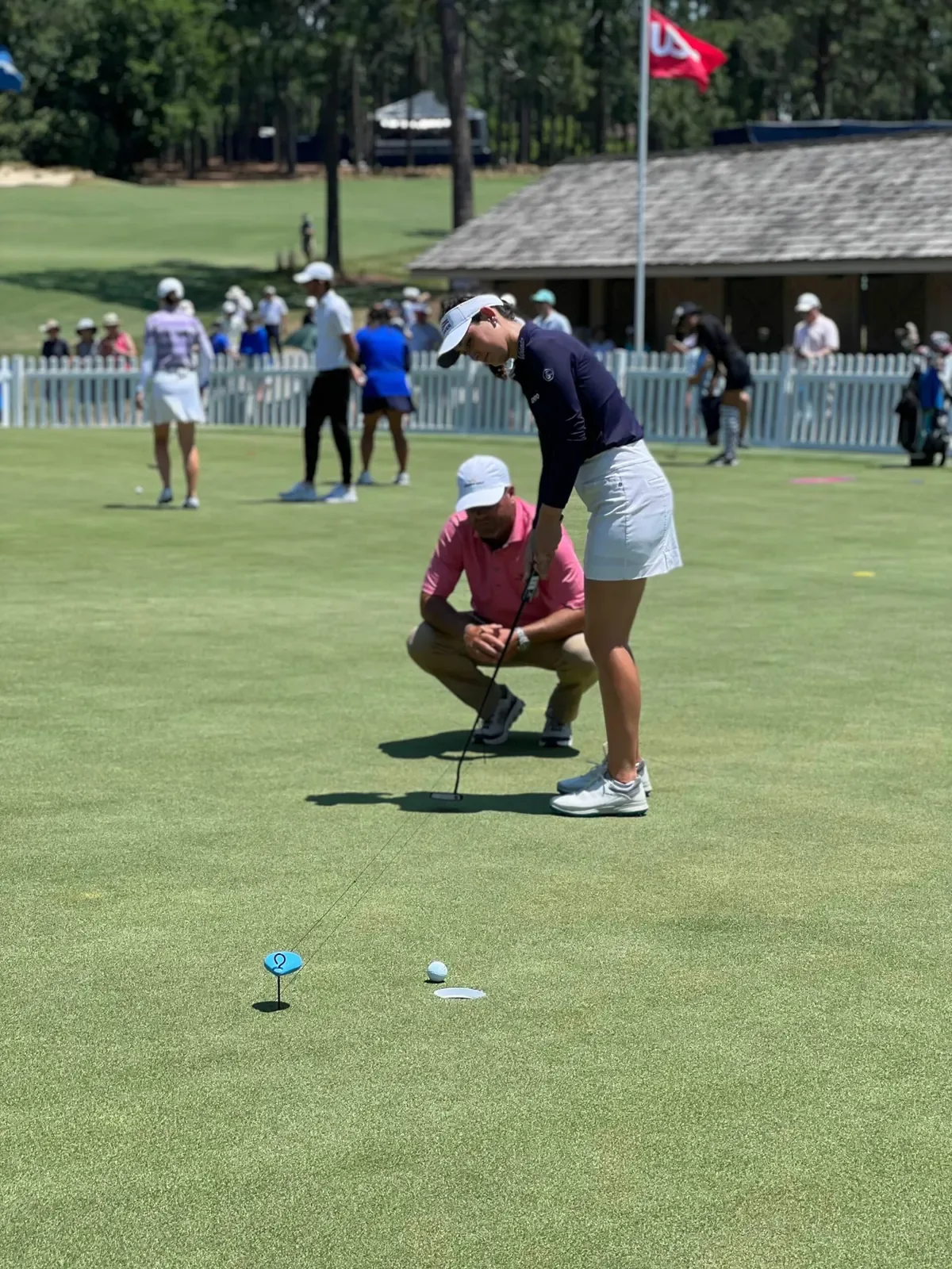 A coach in a pink polo crouches beside a player lining up a putt on a practice green