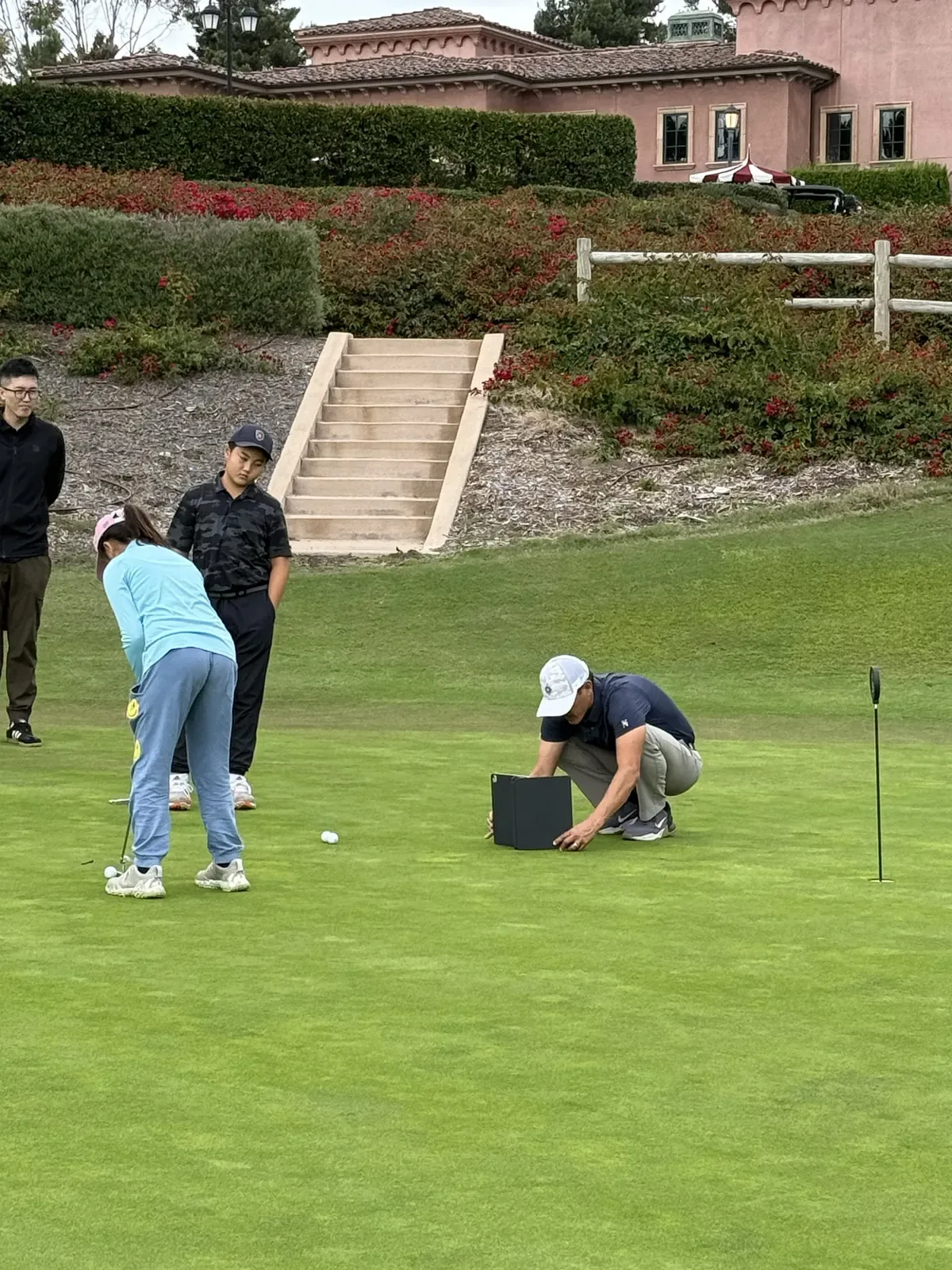 A coach crouches with a tablet on a practice green while a junior putts and others watch
