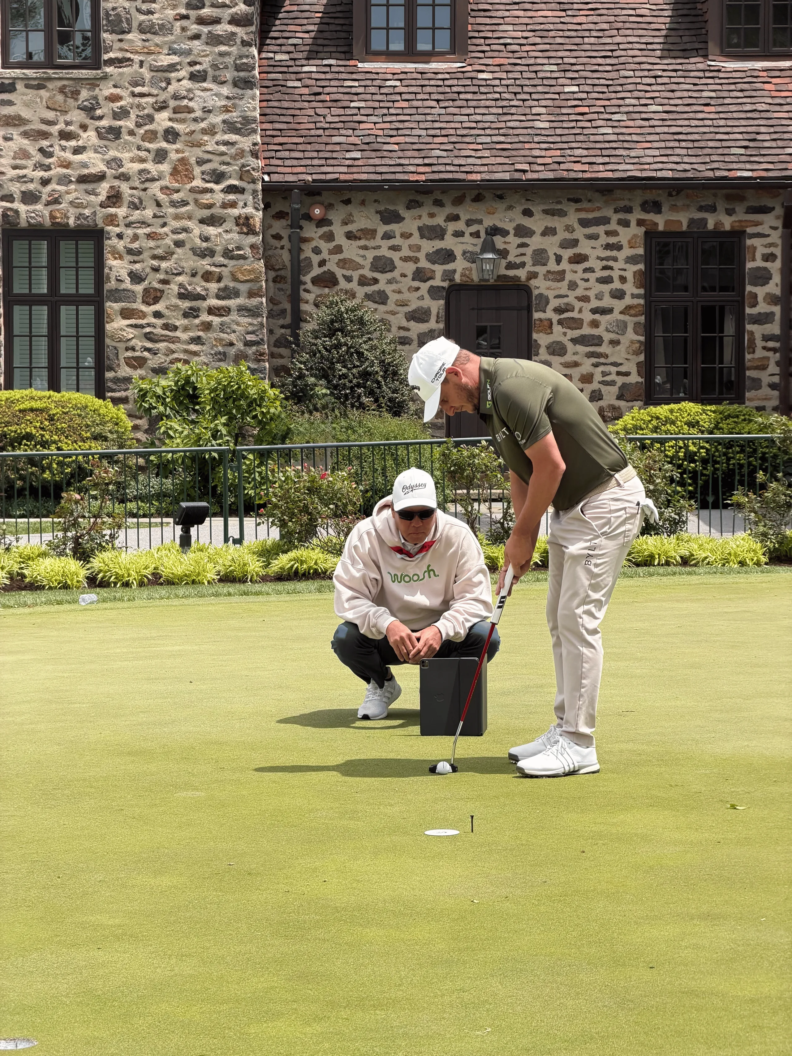 Derek Uyeda crouches behind a player lining up a putt on a practice green in front of a stone clubhouse