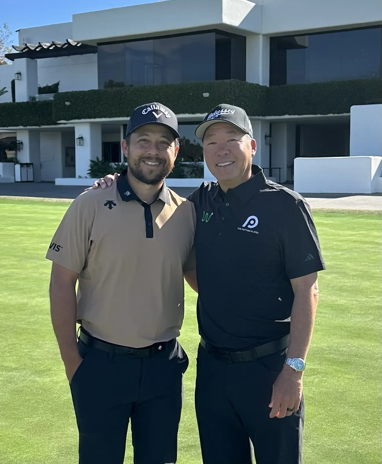 Derek Uyeda and Xander Schauffele stand together with arms around shoulders on a practice green in front of a clubhouse