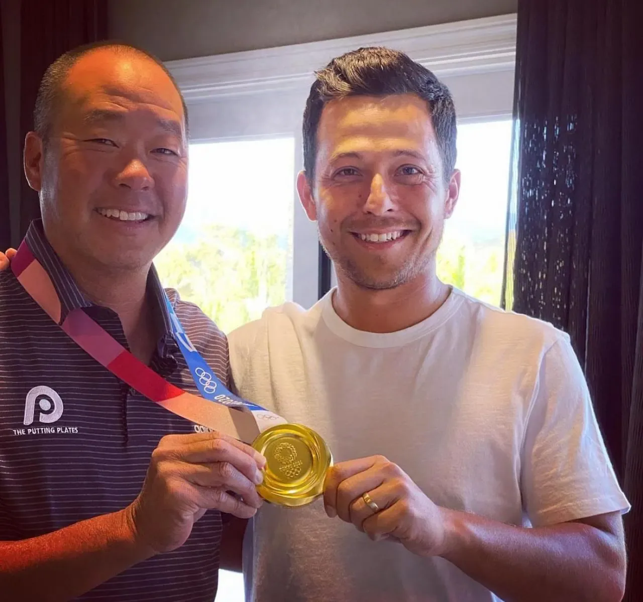 Two men smile indoors while holding a gold Olympic medal on its ribbon between them