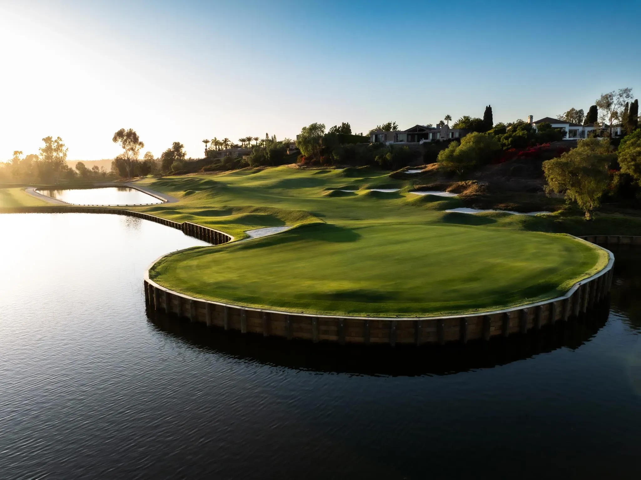 Island putting green ringed by a lake at sunset, with bunkers and homes on the hillside behind