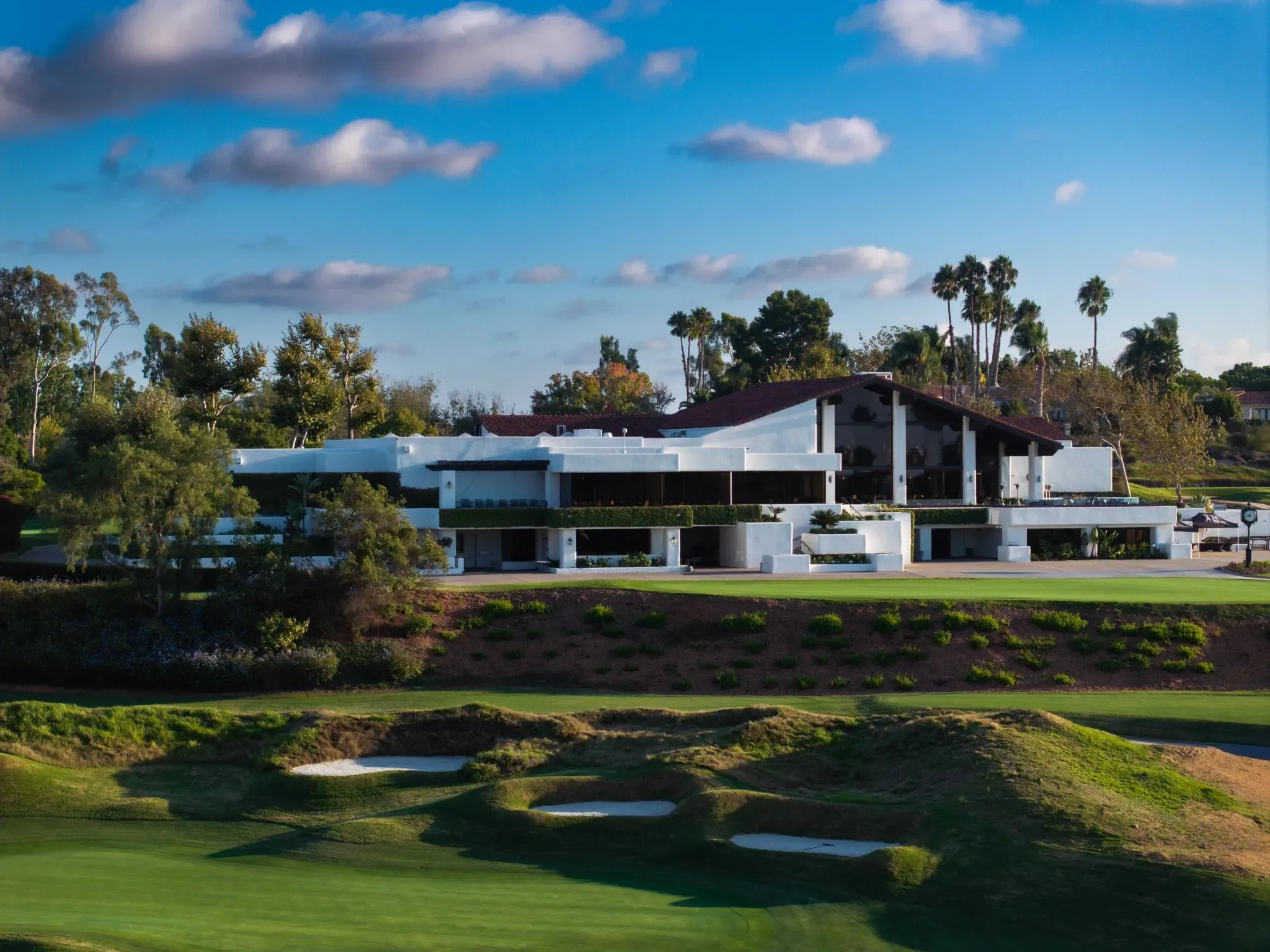 White modernist clubhouse with palm trees above rolling fairway bunkers under a blue sky with scattered clouds