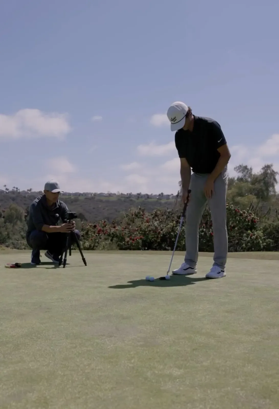 A coach crouches beside a camera on a tripod filming a player putting on a green, with hills behind