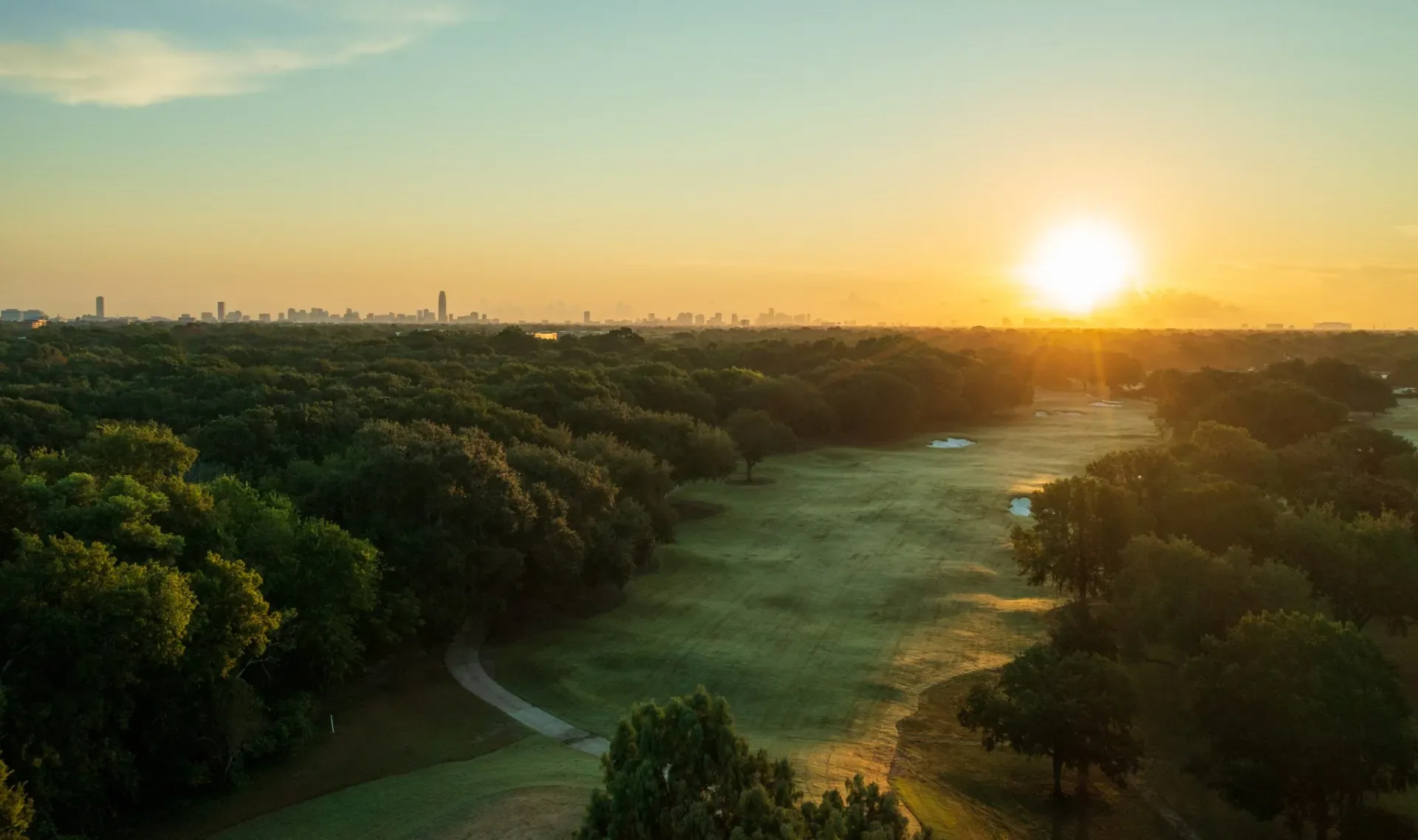 Low sun over a fairway and tree canopy with a distant city skyline at BraeBurn Country Club, Houston