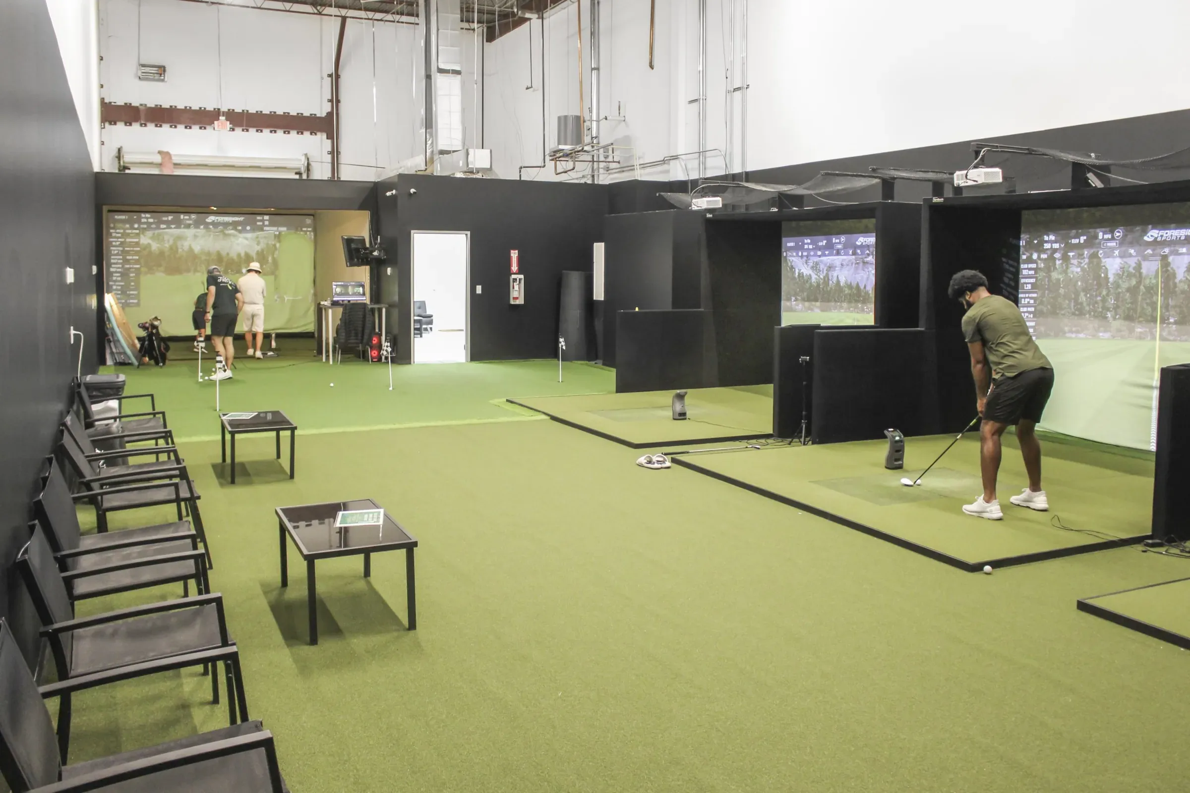 A golfer hits into a simulator screen in a large indoor studio with turf, seating and more bays behind