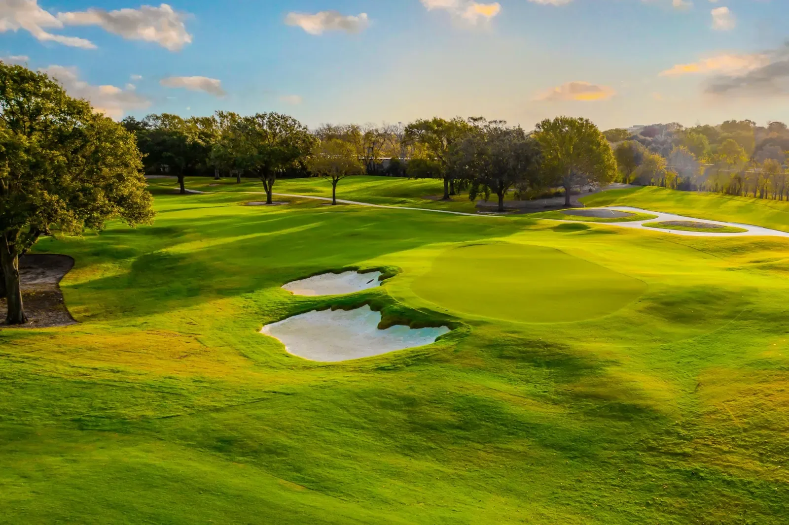Low sun over a green with two bunkers and mature oak trees at BraeBurn Country Club, Houston