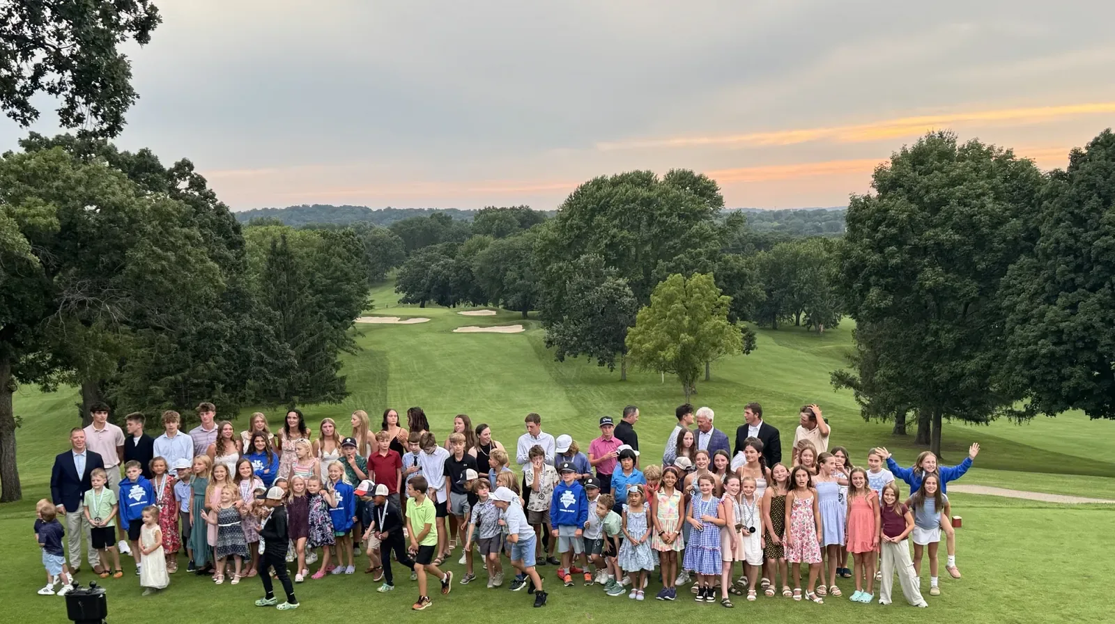 Large group of junior golfers and adults gathered on a tee box with a tree-lined fairway behind them, Balance & Believe Golf.