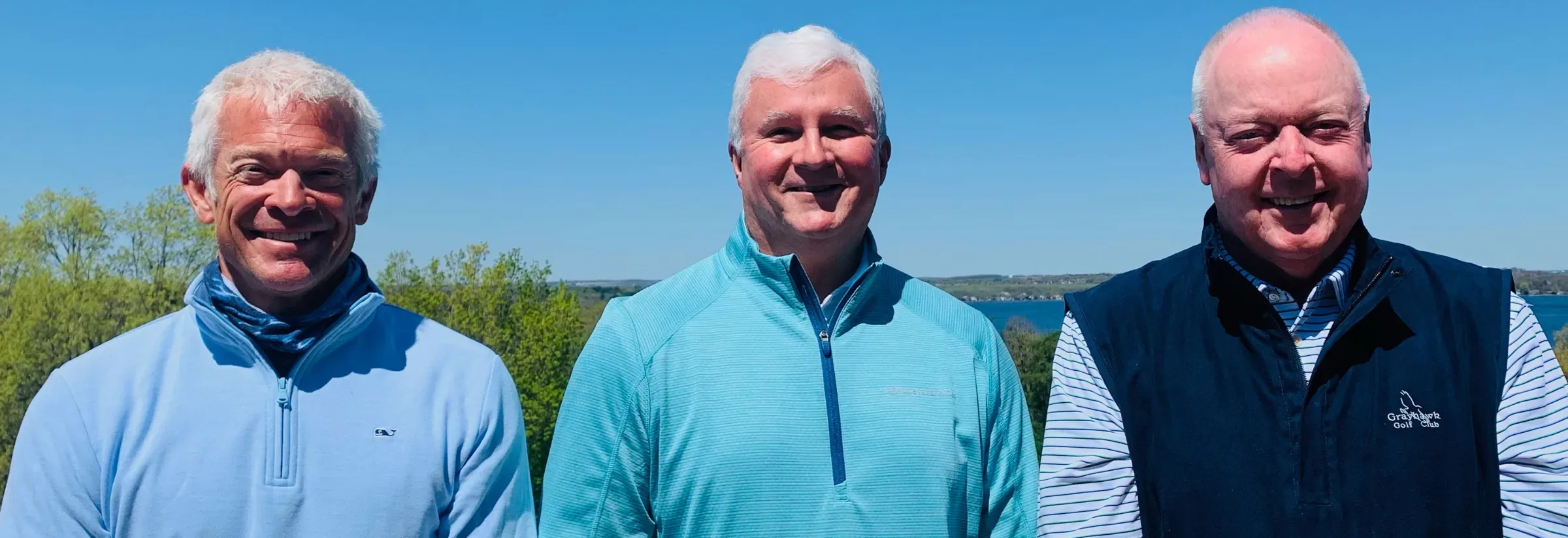 Three men in light blue and navy golf pullovers smiling outdoors with a lake and trees behind them