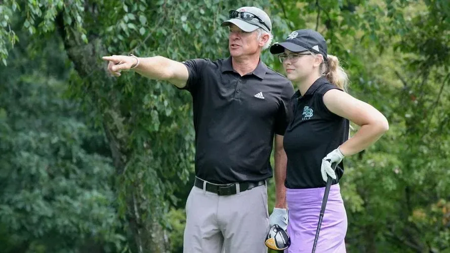 Derek Schnarr in a black polo points into the distance while coaching a golfer on a tree-lined tee.