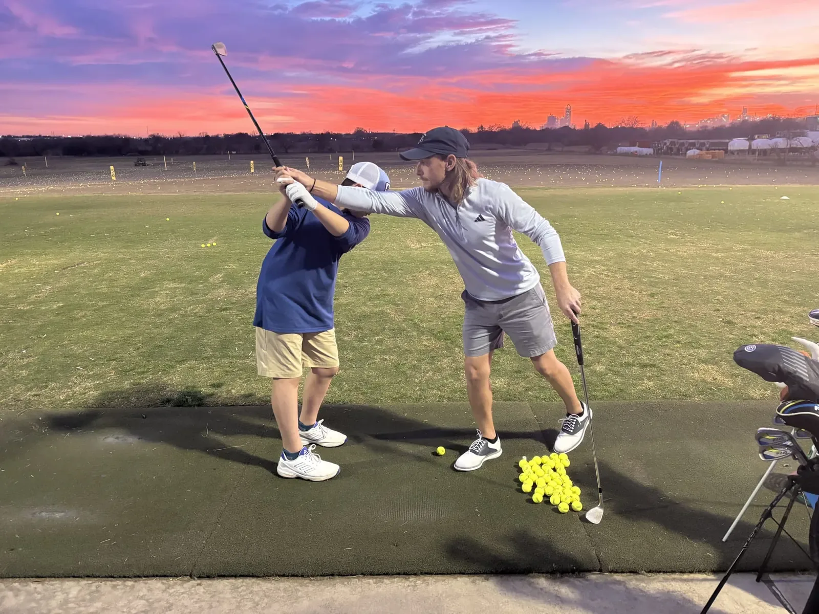 Instructor guiding a young golfer's swing on a hitting mat at sunset, David Ogrin Golf Academy driving range