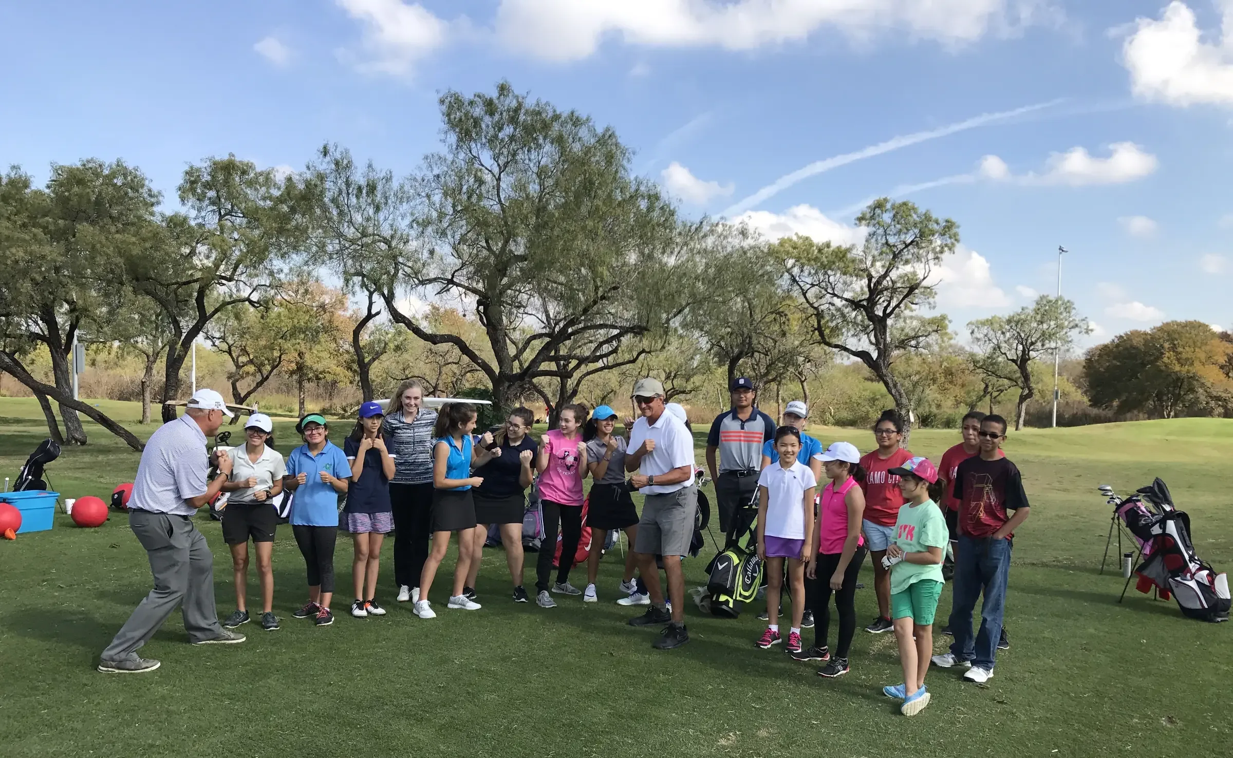 Two instructors and a group of junior golfers raise their fists in a warm-up game on a tree-lined fairway