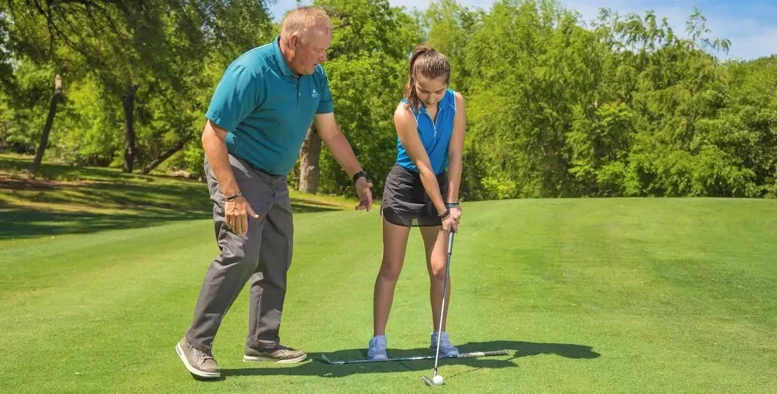 David Ogrin coaching a young golfer at address on a fairway, with a club laid on the grass