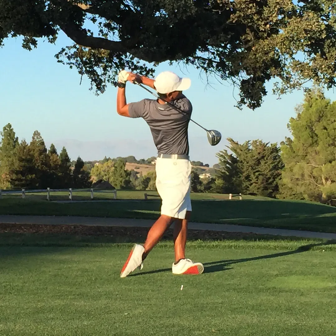 A golfer in a white cap and shorts finishes a driver swing on a tee box under an oak tree