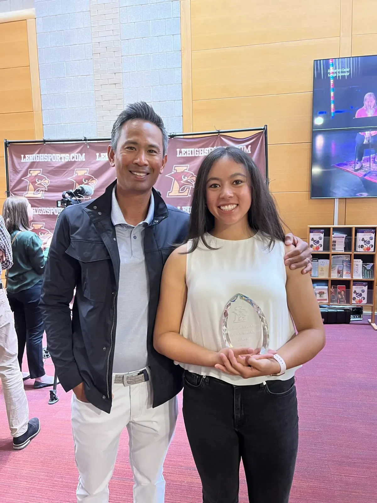 Roger Pineda poses with a young woman holding a crystal award in front of a Lehigh Sports banner
