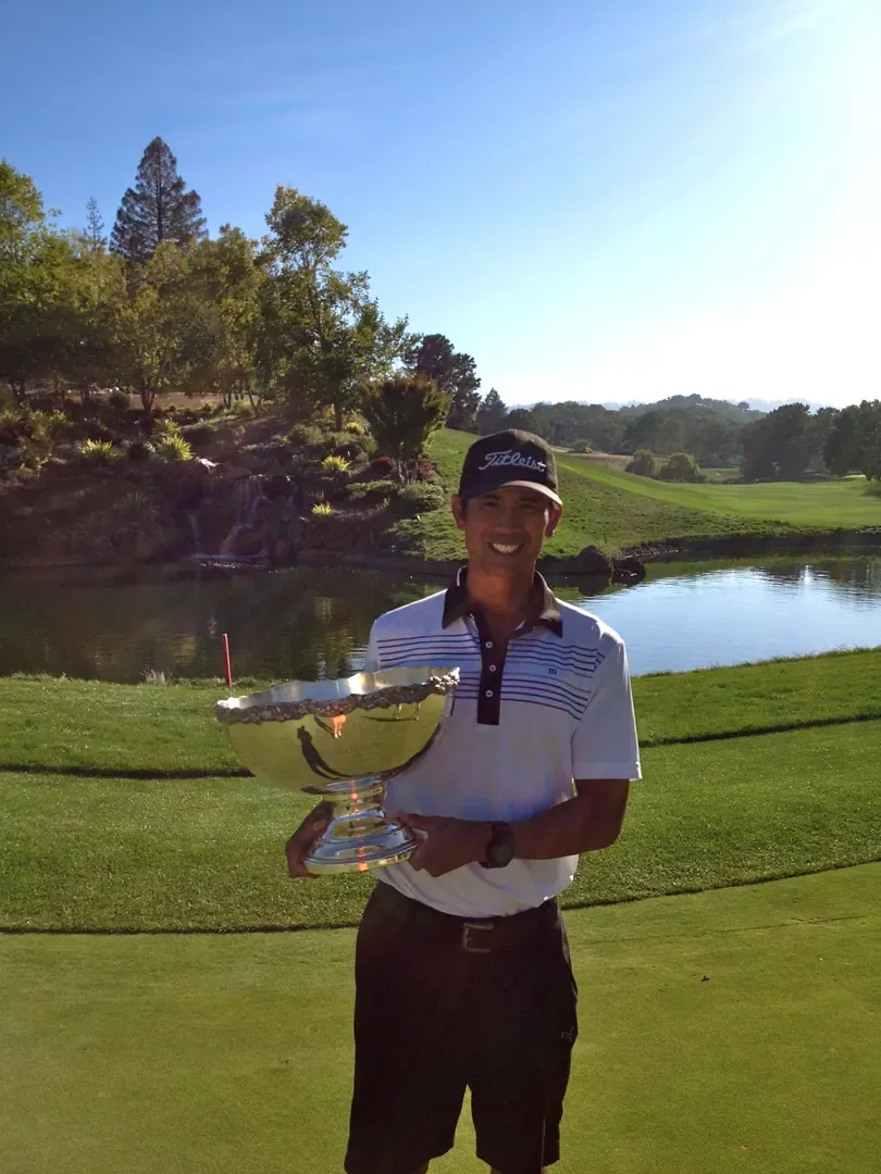 Roger Pineda smiles holding a large silver trophy bowl beside a pond with a small waterfall on a golf course