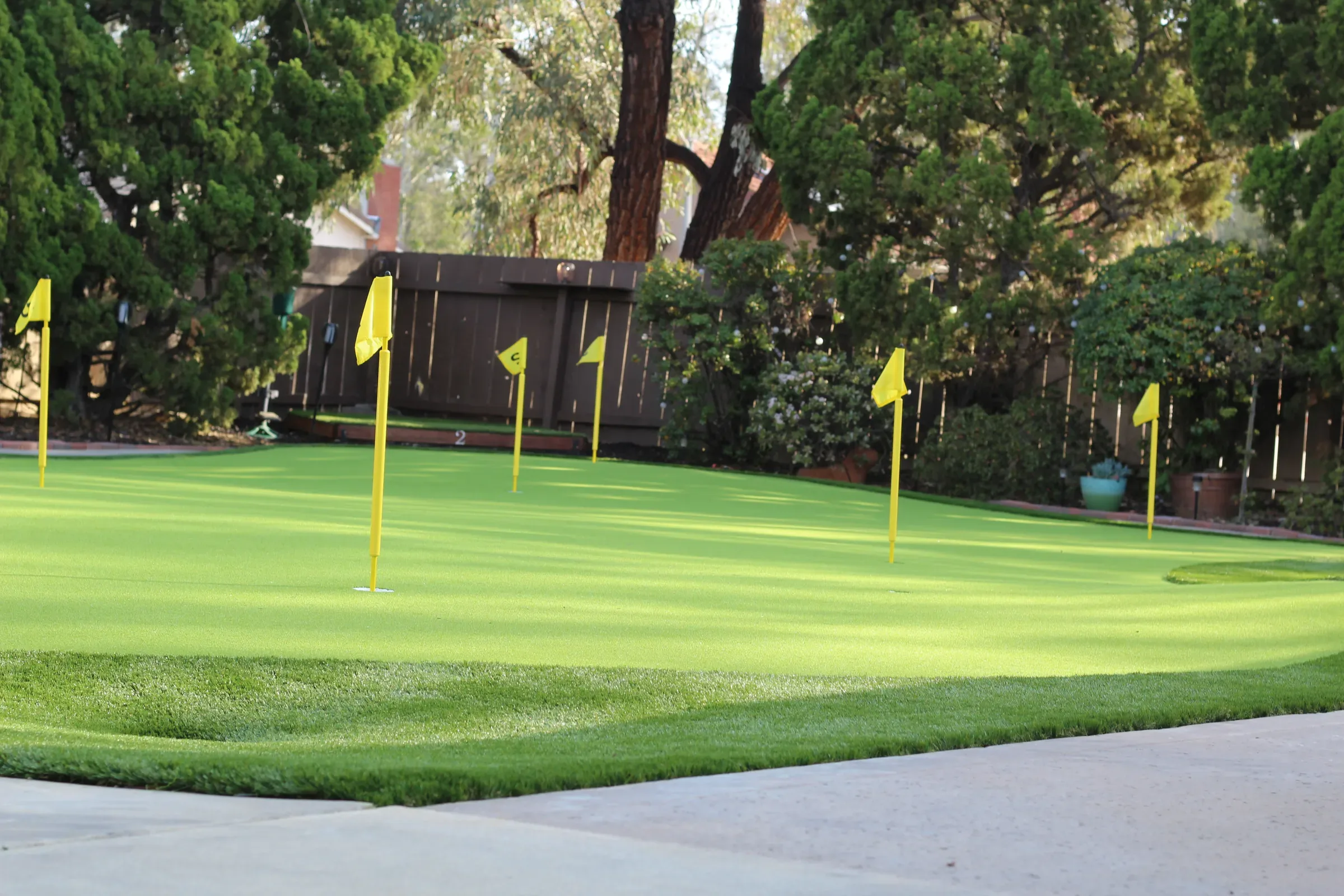 Several yellow flags dot a putting green in front of shrubs, a wooden fence and tall trees