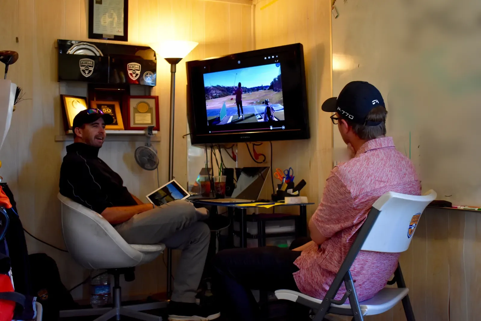 A coach with a tablet and a student watch swing video on a wall-mounted screen in a small lesson room