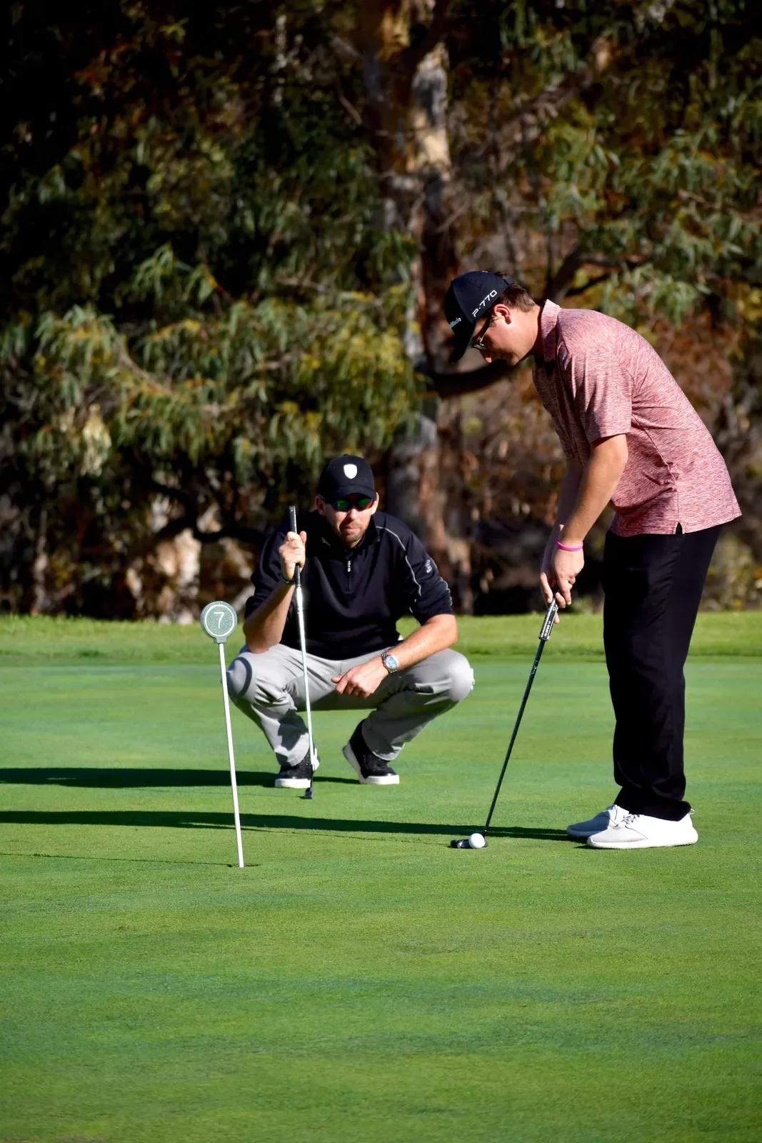 A coach crouches beside a numbered flagstick reading a putt while a golfer putts on a green