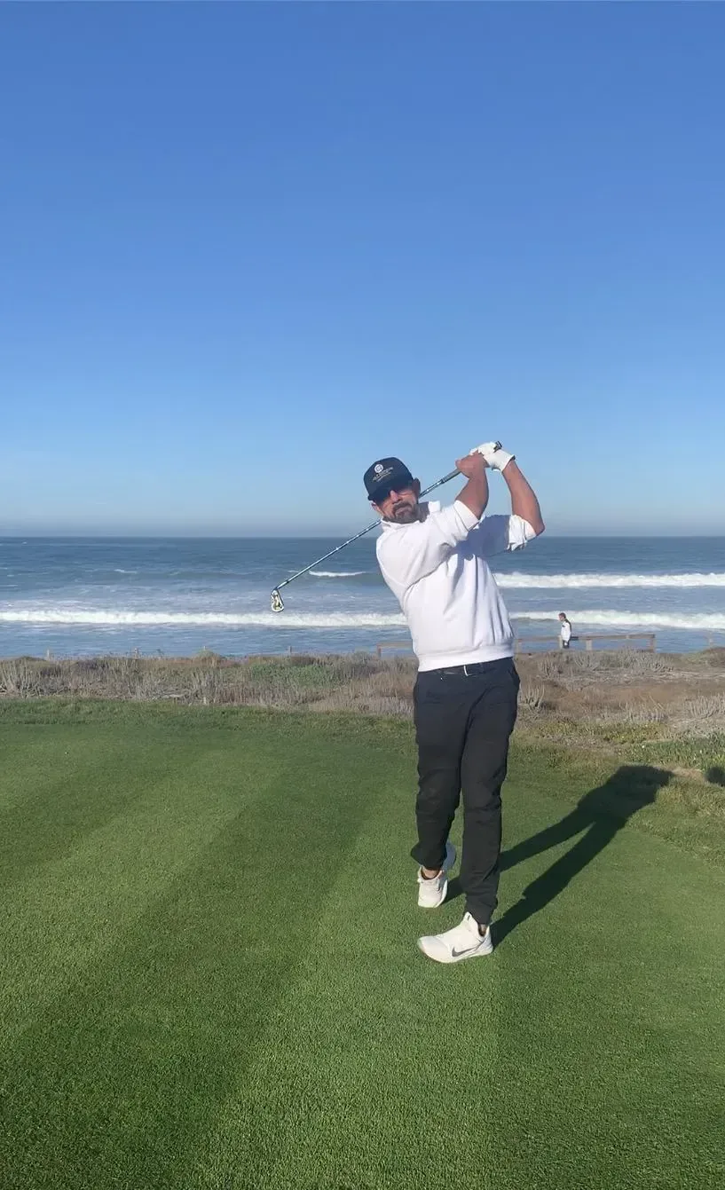 Matt Soza, head professional at The Golf Garage, finishing a swing on a coastal tee with the ocean behind him.
