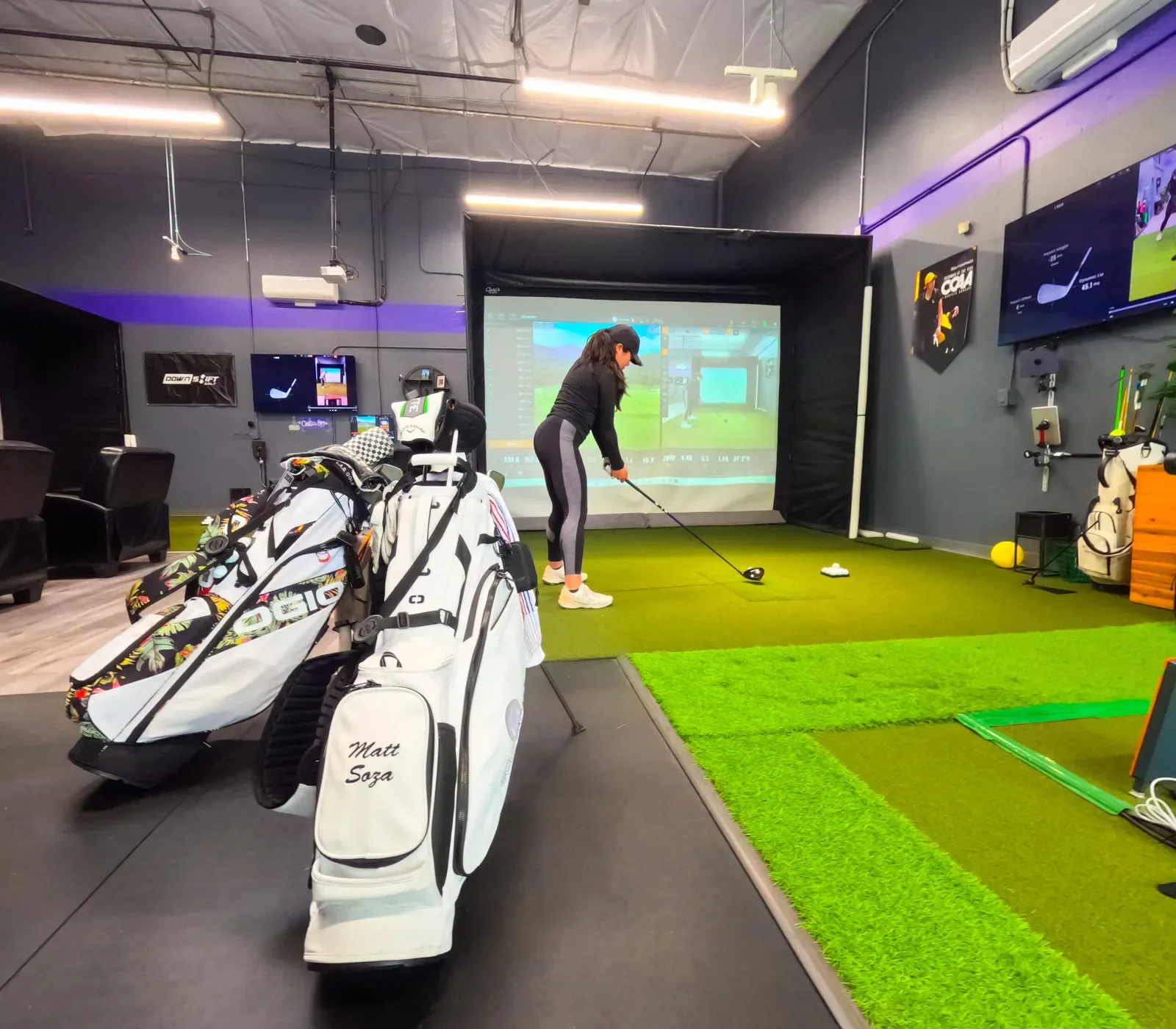 Golfer hits a driver into a simulator screen with two golf bags on the mat, The Golf Garage, San Jose