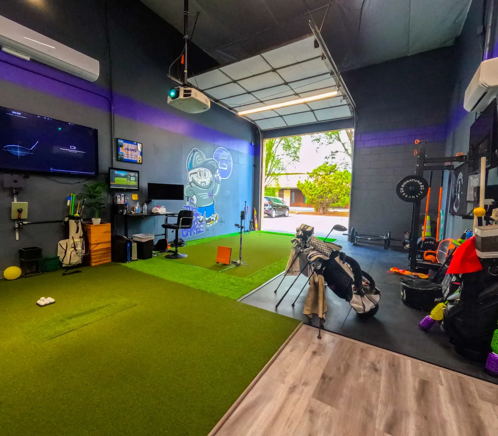 Turf hitting area, monitors and a strength rack beside an open roll-up door at The Golf Garage in San Jose.